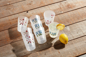 Assorted white plastic cups with blue and green "GO JUMP IN THE LAKE" and red and brown oar designs scattered on a wooden surface with a lemon wedge and a cup of lemonade.