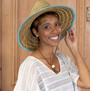 A woman smiles wearing a woven Havana Sun Hat with an Aruba blue ribbon around the brim, complementing her joyful expression and chic, airy attire. The hat’s natural straw color and wide brim offer a classic summer look.