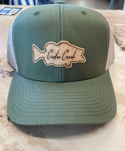 Imagine a trucker hat with moss green front panels and a breathable white mesh back, adorned with a distinctive cork patch shaped like a bass fish and engraved with 