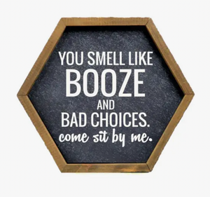 A hexagon-shaped bar sign with a rustic wooden frame and a black background, featuring bold white text that says "You smell like BOOZE and BAD CHOICES. come sit by me.", inviting a humorous and welcoming atmosphere.