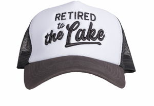 A trucker hat with a white front and dark gray mesh back, embroidered with "RETIRED to the Lake" in stylized black script. The hat embodies a casual yet proud declaration of a relaxed, lakeside retirement lifestyle.