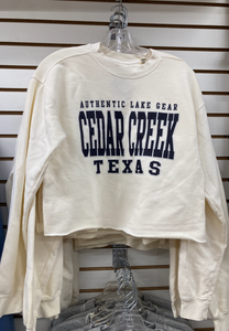 A close-up of a hanging ivory-colored fleece crew sweatshirt with 'AUTHENTIC LAKE GEAR CEDAR CREEK TEXAS' printed in navy blue, displayed against a wooden slatwall background.