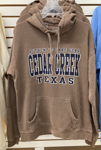 A plush ringspun fleece hoodie in a neutral brown shade with bold "Authentic Lake Gear CEDAR CREEK TEXAS" lettering across the chest, displayed on a hanger, offering a look of classic comfort.