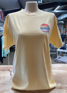 Front view of a butter-yellow ringspun cotton t-shirt on a mannequin, featuring a small 'Cedar Creek Lake Life' logo on the left chest area, in a store environment.