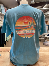 Load image into Gallery viewer, Rear view of a teal ringspun cotton t-shirt displayed on a mannequin with a large &#39;Cedar Creek Lake Life&#39; logo centered on the back, featuring a sunset, trees, and paddles, in a shop setting.
