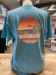 Rear view of a teal ringspun cotton t-shirt displayed on a mannequin with a large 'Cedar Creek Lake Life' logo centered on the back, featuring a sunset, trees, and paddles, in a shop setting.