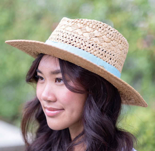 A woman wearing a woven straw sun hat that has an Aruba blue ribbon around the brim, complementing her serene expression and natural backdrop. The hat casts a soft shadow on her face, highlighting her gentle smile.