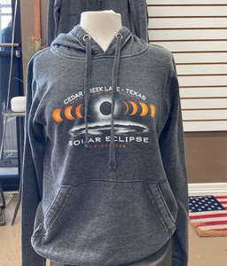 A mannequin wears a charcoal gray fleece hoodie featuring a solar eclipse graphic with phases of the moon and "CEDAR CREEK LAKE - TEXAS SOLAR ECLIPSE 04/08/2024" text, displayed in a store with an American flag in the background, evoking a sense of anticipation for the astronomical event.