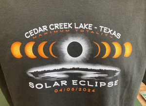Close-up of a dark gray t-shirt featuring an intricate solar eclipse design with the moon covering the sun, accompanied by text 'CEDAR CREEK LAKE - TEXAS MAXIMUM TOTALITY SOLAR ECLIPSE 04/08/2024'.