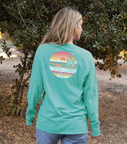 A woman in a Caribbean blue long-sleeved ringspun cotton t-shirt looks over her shoulder, the 'Cedar Creek Lake Life' logo prominent on her back against an outdoor, leafy background.