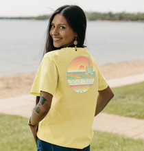 Load image into Gallery viewer, A woman with long dark hair, smiling over her shoulder, wearing a butter-yellow t-shirt with a &#39;Cedar Creek Lake Life&#39; logo on the back, standing by the lake shore.