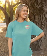 Load image into Gallery viewer, A smiling woman outdoors wearing a teal ringspun cotton t-shirt with a small &#39;Cedar Creek Lake Life&#39; logo on the left chest area, backlit by a sunlit tree background.