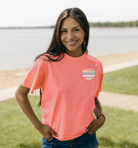 A person standing by the lake wearing a neon coral t-shirt with the Cedar Creek Lake Life logo on the chest.