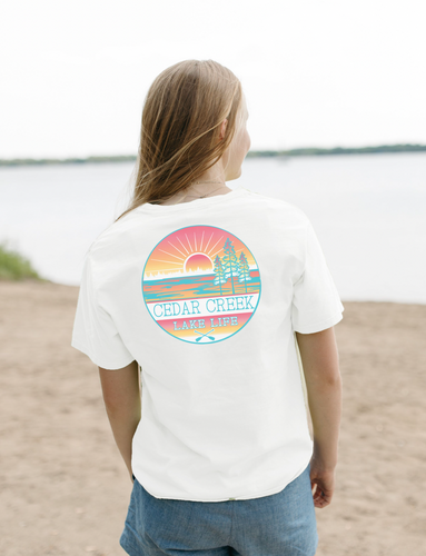 A person facing a lake, wearing a white tee with a colorful Cedar Creek Lake Life logo on the back.