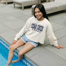 Load image into Gallery viewer, A woman sits by a pool, wearing an ivory cropped ringspun fleece crew with &#39;Cedar Creek Texas&#39; in navy, radiating relaxed lake vibes.