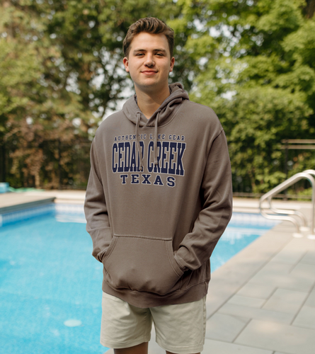 A young man stands confidently poolside, wearing a Cedar Creek Texas ringspun fleece hoodie in a deep taupe shade that radiates casual style and lakeside pride.