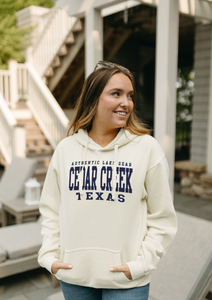 A woman stands on a porch, smiling and wearing a cream-colored fleece hoodie with "Authentic Lake Gear CEDAR CREEK TEXAS" printed in navy blue. The hoodie exudes a relaxed, comfortable style, perfect for a casual lakeside lifestyle.