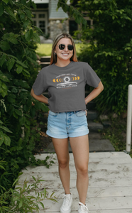 A young woman smiles in a gray cropped overdyed tee with Cedar Creek Lake Solar Eclipse design, paired with denim shorts, in a lush garden setting.