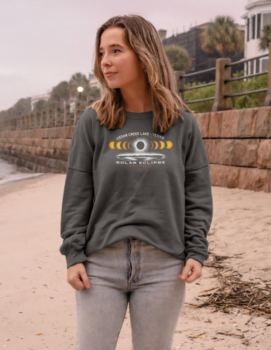 A young woman stands on a beachside walkway, wearing the Cedar Creek Lake Eclipse fleece crewneck in a relaxed fit, evoking the spirit of anticipation for the solar eclipse event.