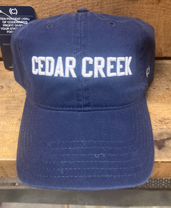 A navy blue baseball cap with "CEDAR CREEK" embroidered in white block letters displayed on a wooden shelf, capturing a casual, outdoor-ready aesthetic. The hat symbolizes a connection to Cedar Creek Lake and a love for the laid-back lake lifestyle.