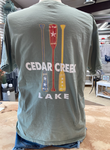 A person stands with their back turned, wearing a sage green, short-sleeved t-shirt with a graphic of three canoe paddles in red, white, and blue, the middle paddle featuring a star design. Above and below the paddles, the words "CEDAR CREEK LAKE" are printed in block letters.