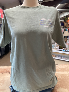 The front of a sage green, short-sleeved t-shirt displayed on a mannequin. A small, faded graphic of crossed canoe paddles with stars and stripes sits on the left chest area, with "CEDAR CREEK LAKE" printed above and below in a semicircle.
