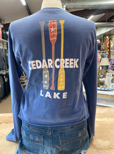 The back of a navy blue cropped ringspun cotton tee, featuring a vertical graphic of three paddles painted in red, white, and yellow with star details, placed above the words 'CEDAR CREEK LAKE' in white block letters.