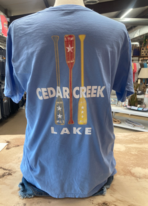 A cornflower blue ringspun cotton t-shirt displayed on a mannequin, showcasing the back design with three colorful paddles and the 'CEDAR CREEK LAKE' print in bold white letters.
