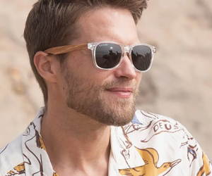 Man in patterned shirt smiling and wearing Blue Planet eco-friendly crystal-clear sunglasses with zebra wood arms and polarized lenses for stylish sun protection.