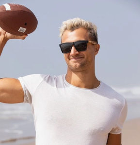 Smiling man on beach wearing eco-friendly bamboo temple sunglasses with polarized lenses, casually holding a football, symbolizing active lifestyle and sustainable fashion.