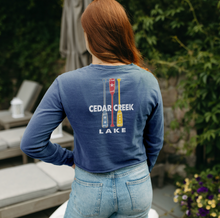 Load image into Gallery viewer, A woman stands outdoors, wearing a navy blue Cedar Creek Lake paddles cropped tee, showing the full back design with paddles graphic and lettering against a backdrop of lush greenery.