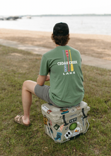 Load image into Gallery viewer, A person wearing a sage green t-shirt with the &quot;CEDAR CREEK LAKE&quot; paddles graphic on the back sits on a sticker-covered cooler by the lakeshore, facing the water. The relaxed pose and scenic backdrop convey a sense of leisure and enjoyment typical of lakeside living.