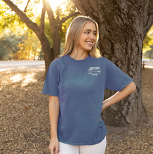 Load image into Gallery viewer, A woman wearing the Cedar Creek Lake Paddles Ringspun Tee in heather green, smiling and standing outdoors with sun  -filtered trees in the background, showcasing the front design with &#39;CEDAR CREEK LAKE&#39; text on the left chest.