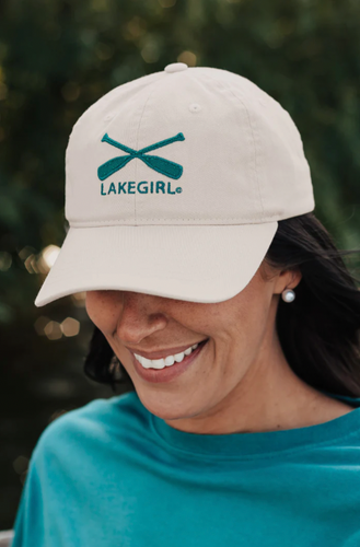 A woman smiling, wearing a cappuccino-colored 'Lake Girl' cap with crossed paddles logo in teal, conveying a joyful lakeside lifestyle.