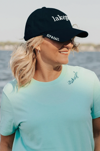 A woman in a light blue t-shirt smiles while wearing a navy "Lakegirl" cap with white lettering and the phrase "SIP & SAIL" on the side, against a serene lakeside backdrop.