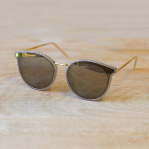 The Canaveral sunglasses rest on a wooden surface, showcasing their modern elegance. These round-framed glasses feature a muted olive acetate upper with sleek gold metal arms. The gray lenses fade subtly into the frames, offering a harmonious and stylish look suitable for a range of personal styles. The understated design of these sunglasses speaks to a contemporary, minimalist aesthetic.