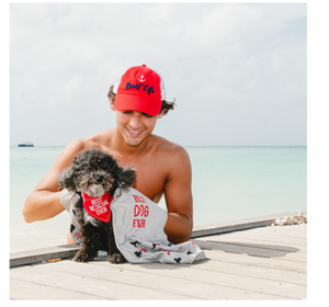 A man holding a small dog, both sporting red gear: the man wears a red mesh hat with the words "Boat Life" and an anchor, and the dog wears a bandana reading "BEST RESCUE EVER." They're enjoying a sunny day by the sea, sharing a bond of companionship and love for the waterfront.