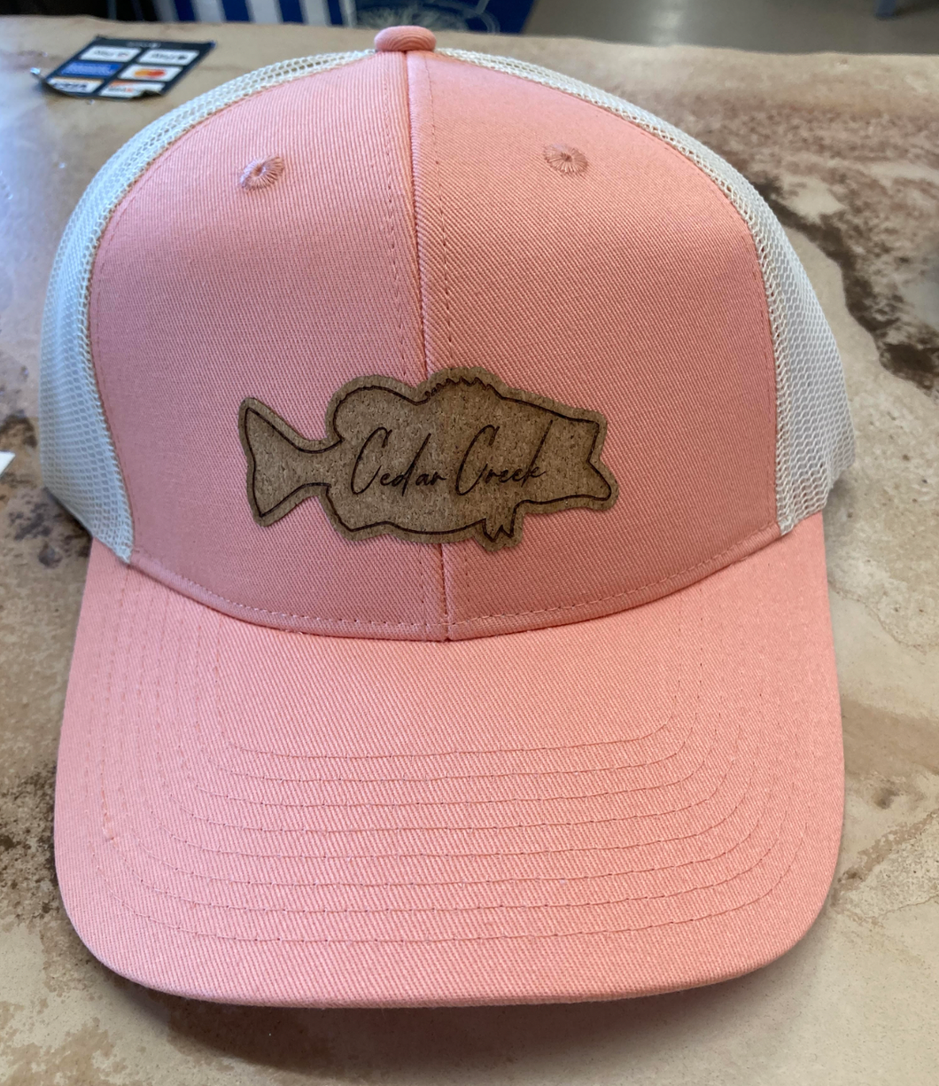 A coral-pink trucker hat featuring a distinctive bass-shaped cork patch with the words 